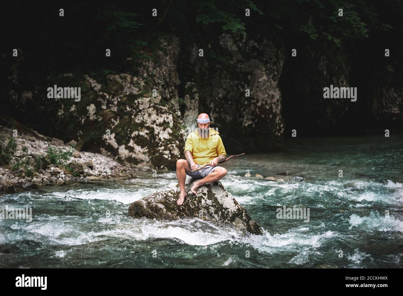 Adventurer with beard sitting in the middle of river on stone and carving on piece of wood Stock Photo