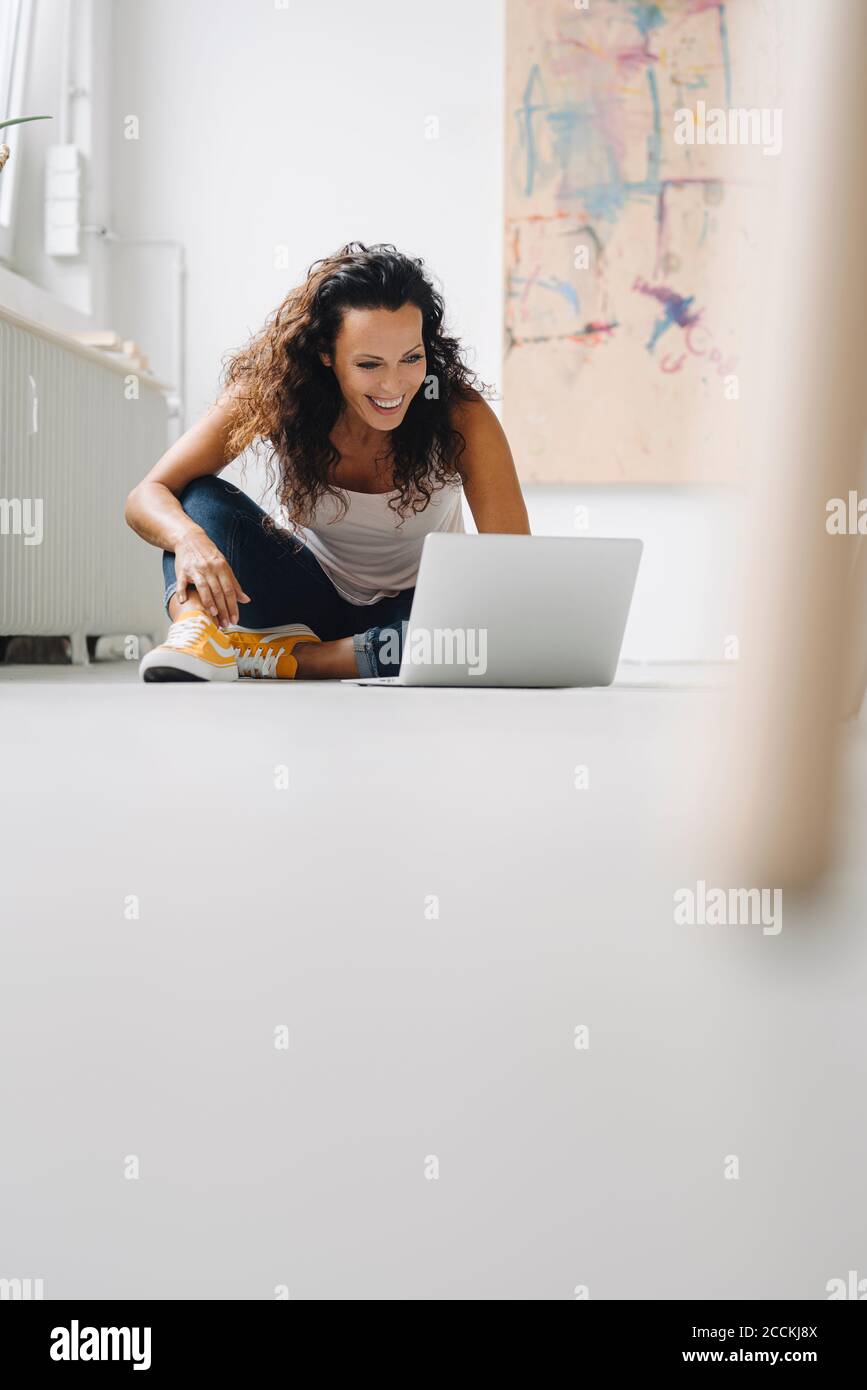 Cheerful mid adult woman using laptop while sitting on floor at home ...
