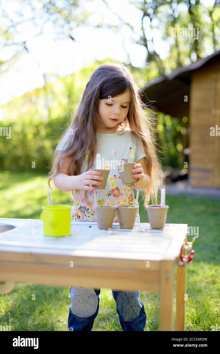 Cute girl planting seeds in small pots while standing at table in yard Stock Photo