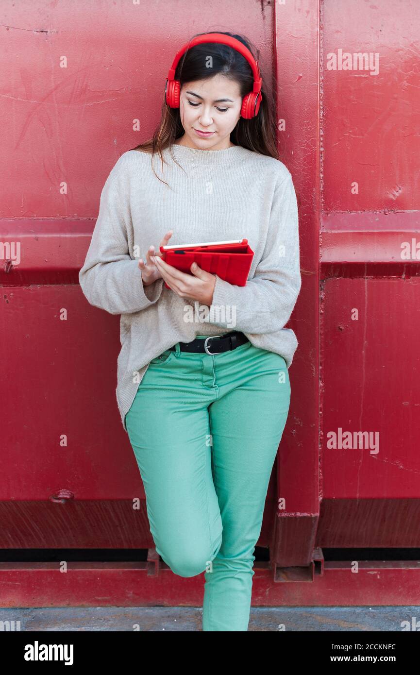 Trendy beautiful woman listening to songs while using digital tablet against red metallic wall Stock Photo