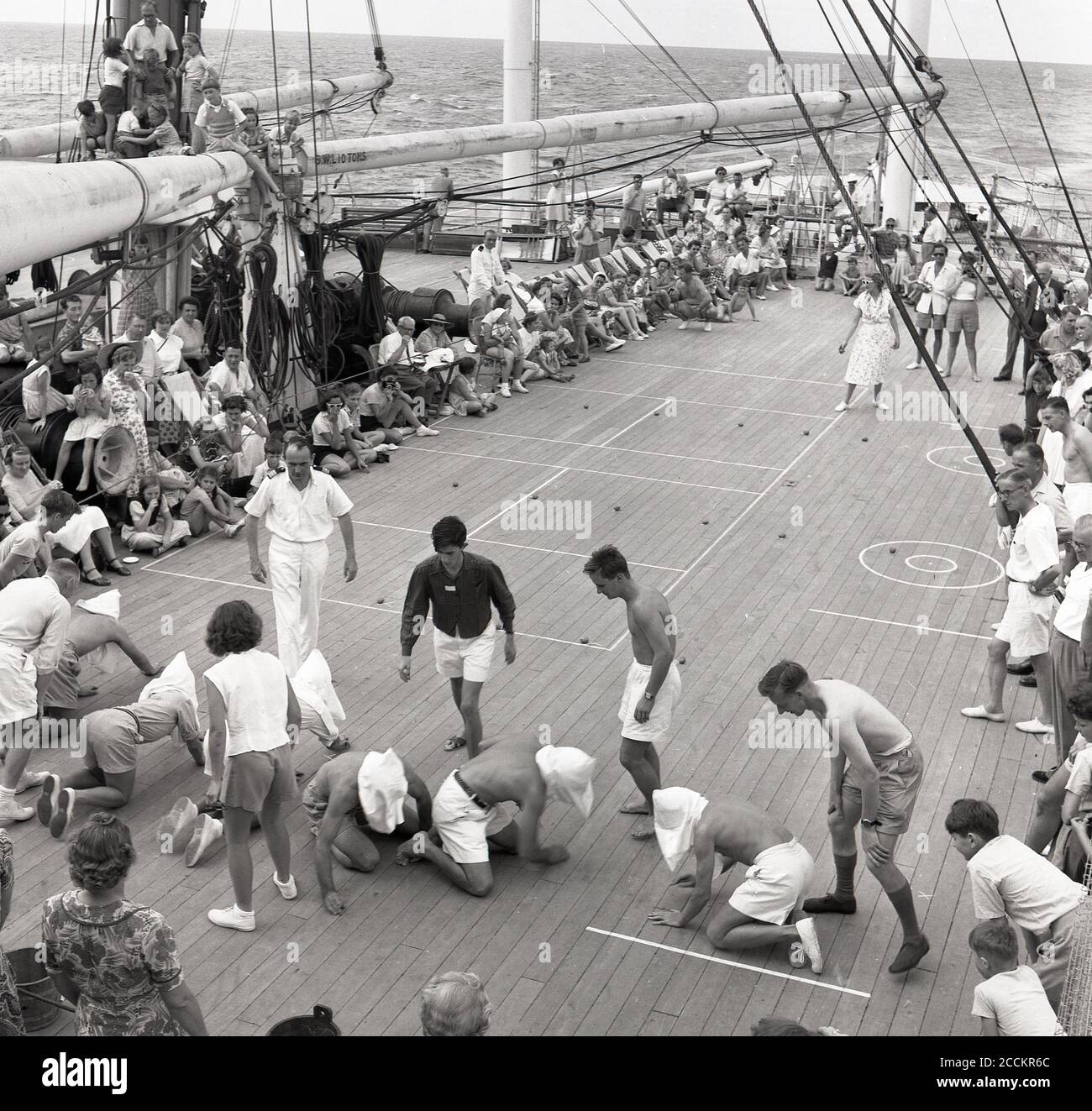 1950s, historical, on-board a Union-Castle steamship headed fot the Cape in South Africa, some male passengers playing a deck game, with sacks over the heads, acting as blindfolds, they have to hunt on their hands and knees searching for hidden items, watched by the ship's other passengers. Stock Photo