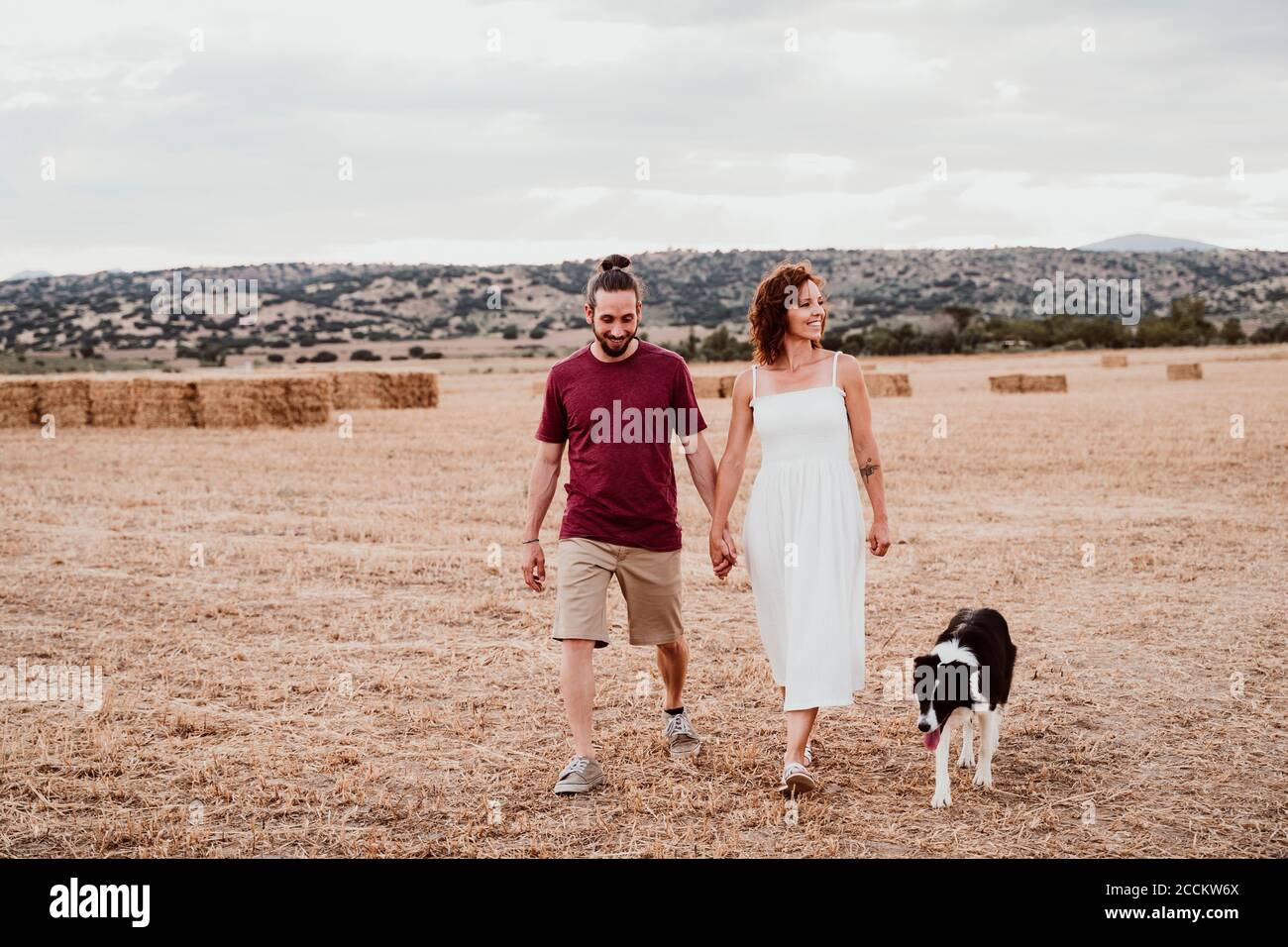 Smiling couple holding hands while walking with dog in field Stock Photo