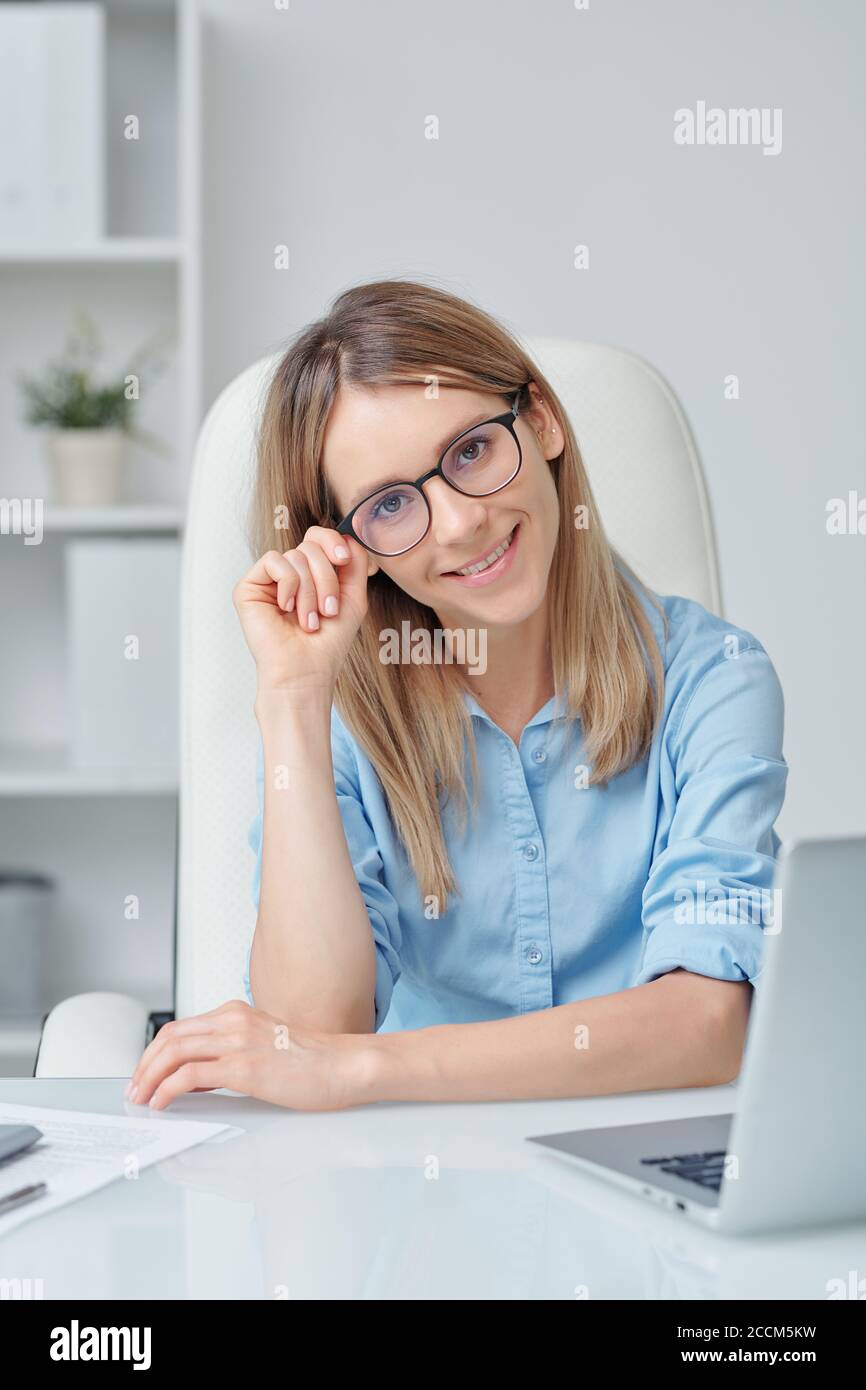 Happy young female office manager in eyeglasses and blue shirt looking at you Stock Photo