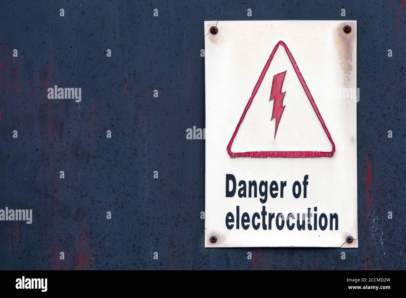 A sign that reads 'Danger of electrocution'. The sign is old and faded. It is on the front of an old black metal door. Stock Photo