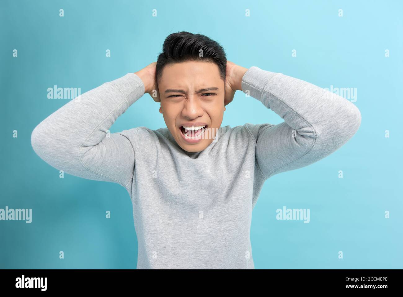 Emotional stress. Frustrated young man holding hands on ears while standing against blue background Stock Photo