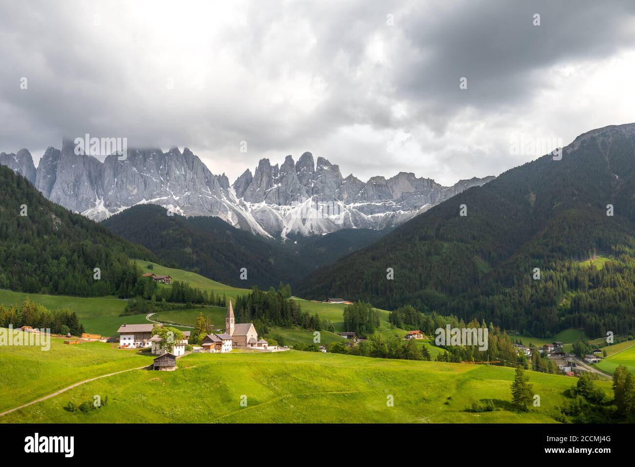 Wide angle view of an Italian valley, with a small church surrounded by meadows and trees in the foreground and a mountain range in the background Stock Photo