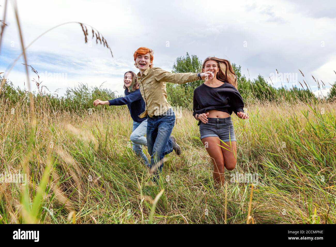Summer holidays vacation happy people concept. Group of three friends boy and two girls running and having fun together outdoors. Picnic with friends on road trip in nature Stock Photo