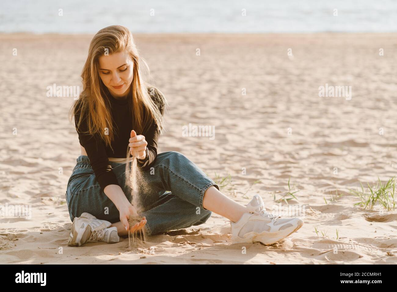 Full length beautiful woman sitting on beach at sunset in evening and pour sand. Slow life, social detox and self-immersion, single. Pretty female with long brown hair in casual clothes. Stock Photo