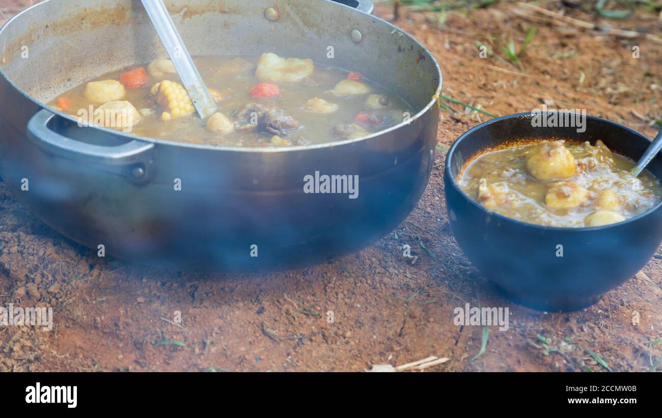 A chicken and dumplin bouillion or soup cooked on a wood fire in the outdoors in a metal pot an a black ceramic bowl resting on the dirt Stock Photo