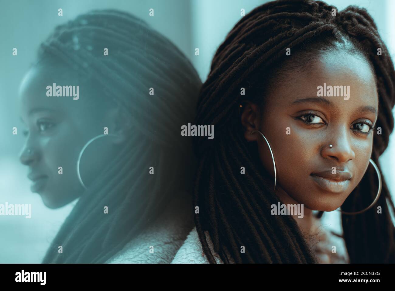 A portrait of admirable young black female from Guinea-Bissau with big earrings, nose piercing, and braided hair, she is leaning against glass wall Stock Photo