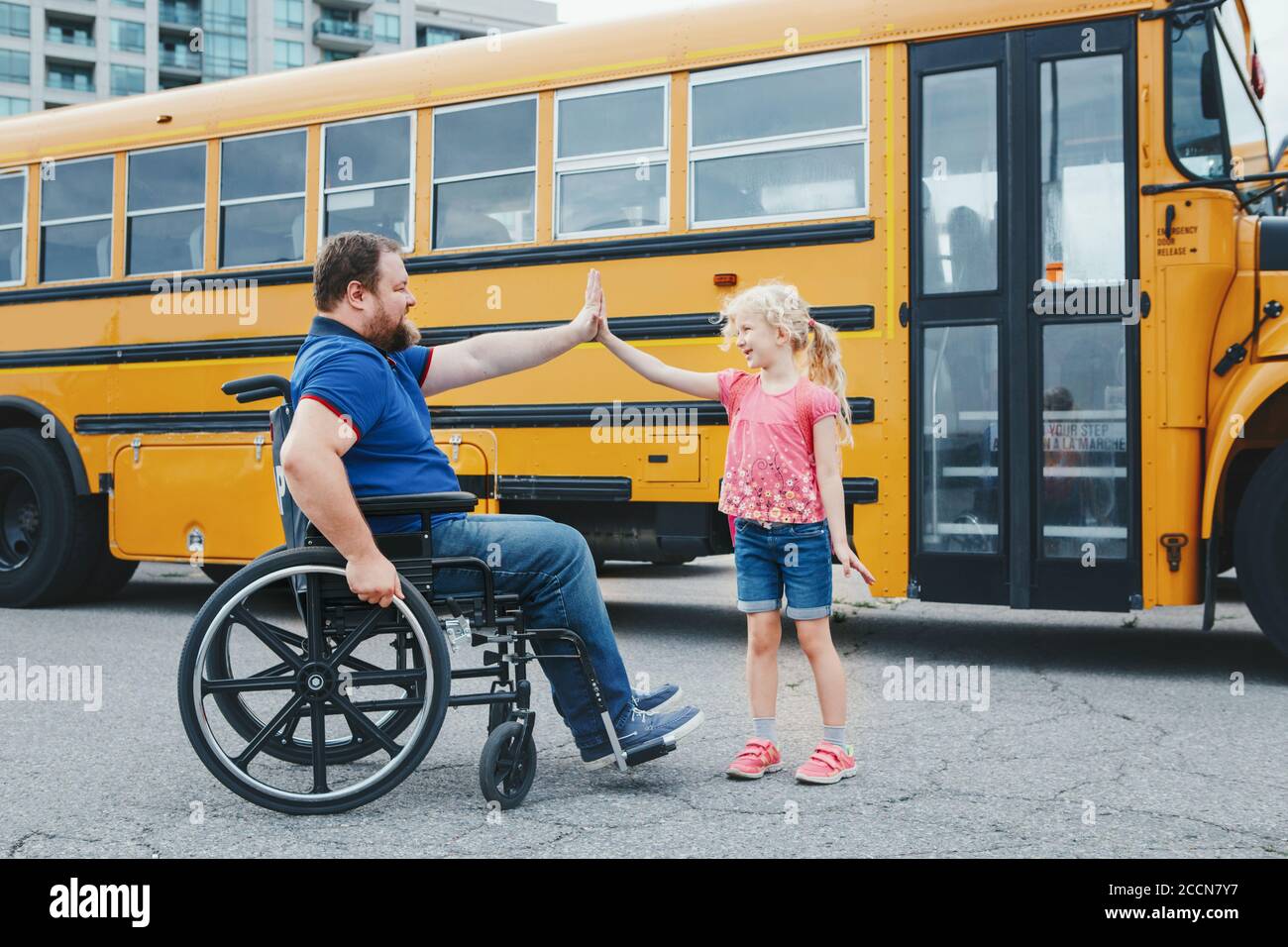 Encourage and support from family members. Disabled Caucasian father on wheelchair with daughter by yellow school bus. Back to school. High five Stock Photo