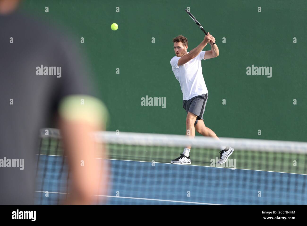 Tennis player man hitting backhand returning ball with racket on green background. Sports men playing together on outdoor court Stock Photo