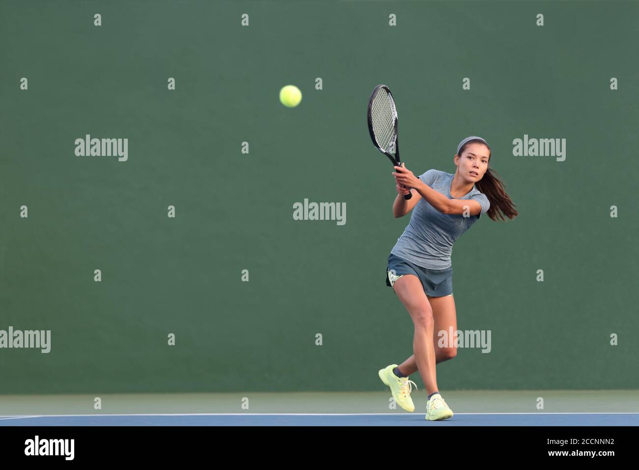 Tennis playing woman hitting ball on green hard court. Asian athlete girl returning serve with racket wearing skort and shoes Stock Photo