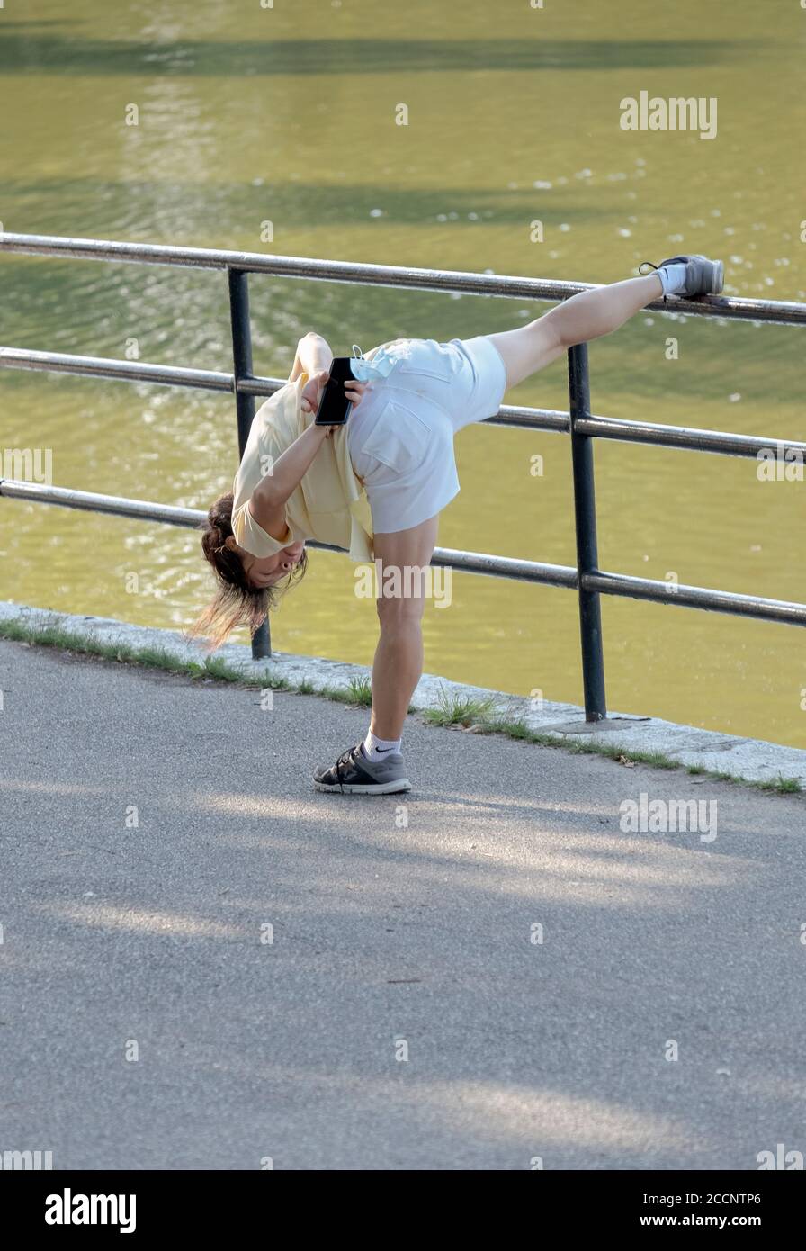 A very limber & nimble Asian American woman holding her mask and cell phone stretches before exercising. In Kissena Park, Flushing, NYCity. Stock Photo