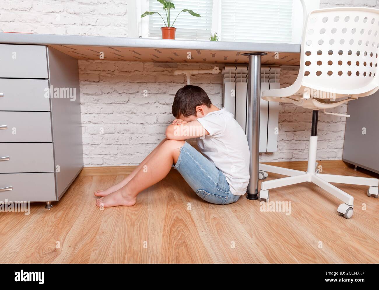 Preteen boy sitting under desk, covering his face with hands at home