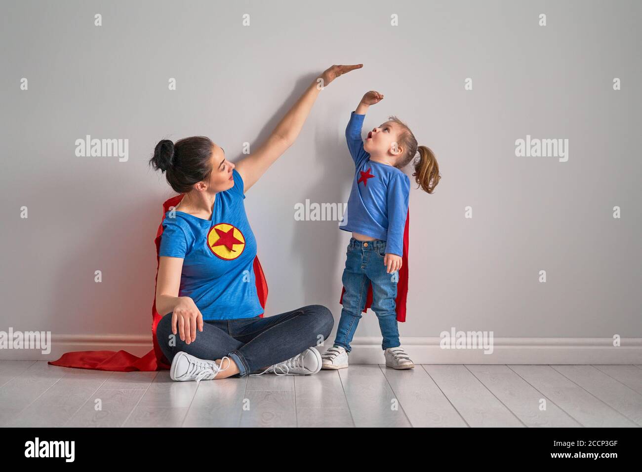 Mother is measuring growth of child daughter near empty wall. Girl in superhero costume. Stock Photo