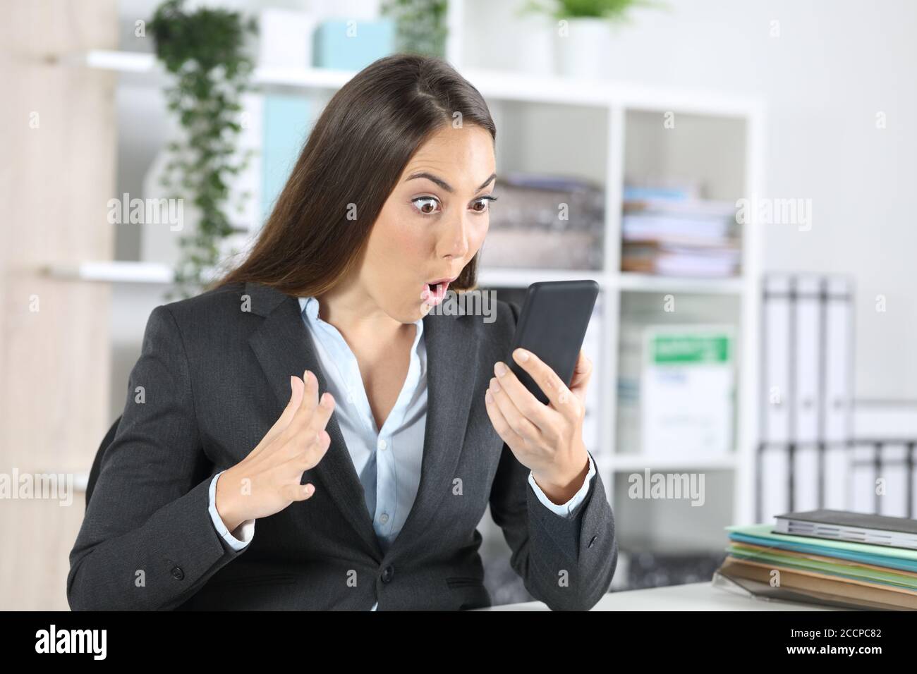 Amazed executive woman checking smart phone sitting on a desk at the office Stock Photo