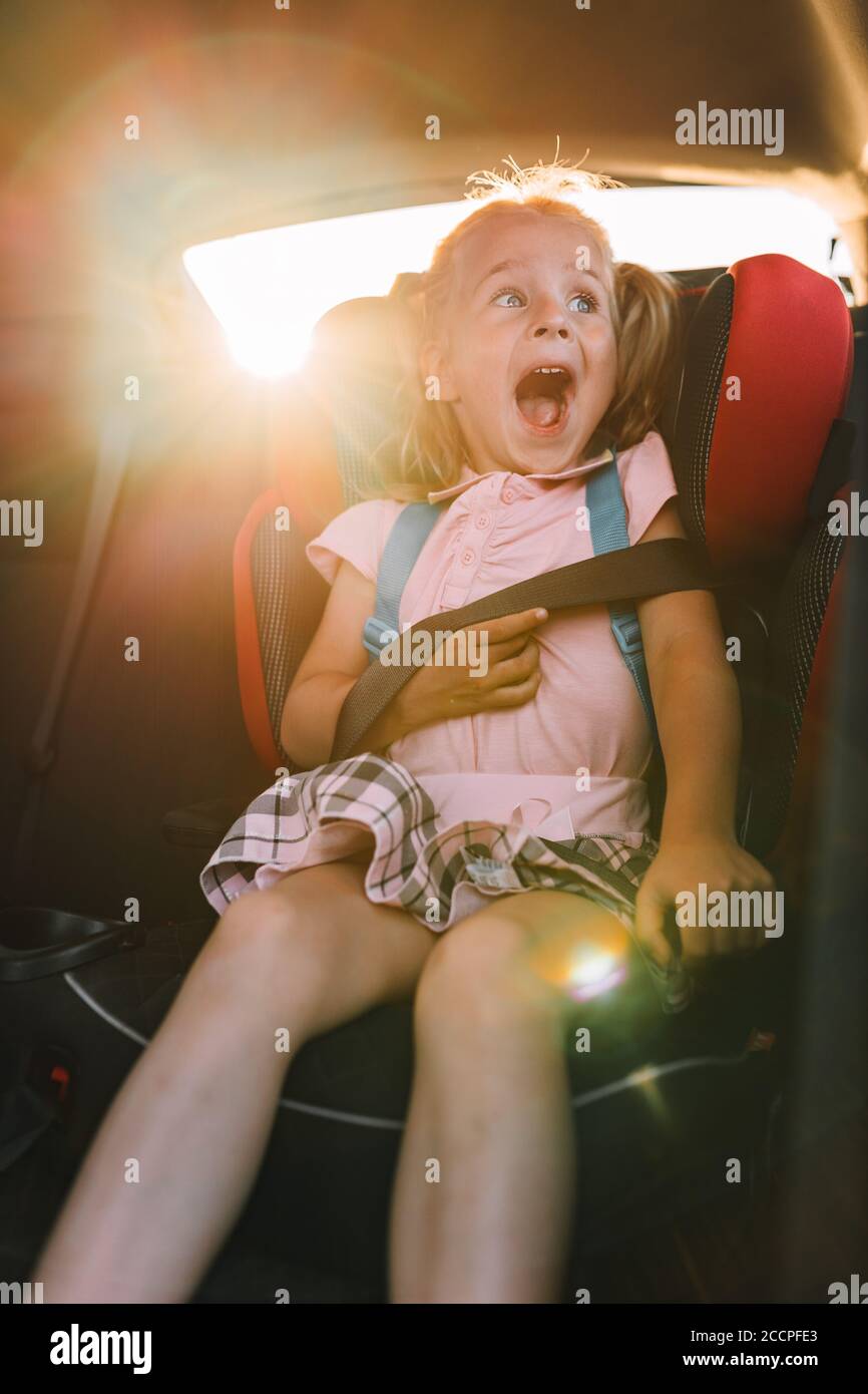 Emotional portrait of a toddler girl sitting safely in car seat with fasten seat belt. Travel with kids, protection on road, vacation, road trip. Stock Photo