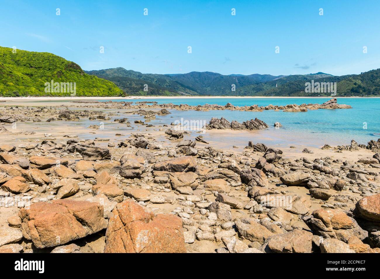 The rocks beach is just next to the shallow and turquoise ocean. In the background lay temperate forests and hills with blue skies. Stock Photo