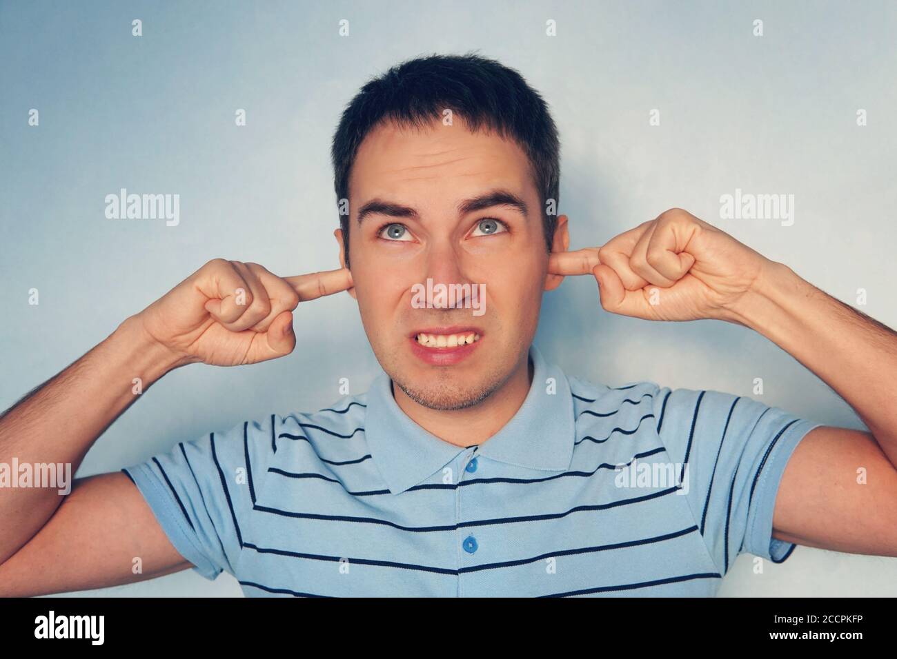 Closeup portrait young, angry, unhappy, stressed man covering his ears, looking up, to say, stop making loud noise it's giving headache, blue Stock Photo