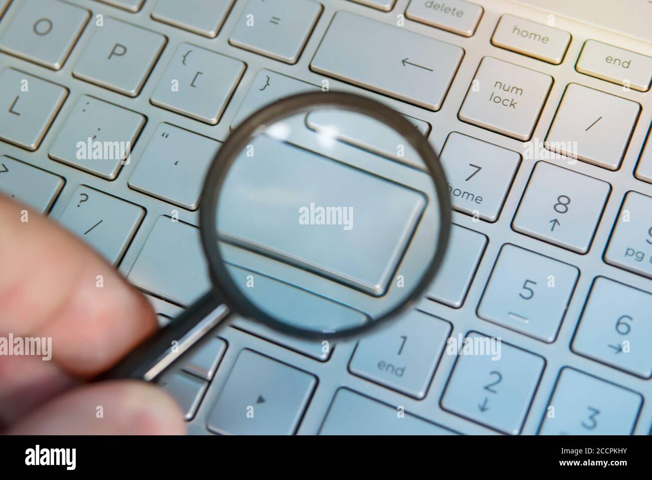 Male hand holds the magnifying glass above the laptop english layout with blank enter key. Your word on a empty computer keypad button. Designer Stock Photo