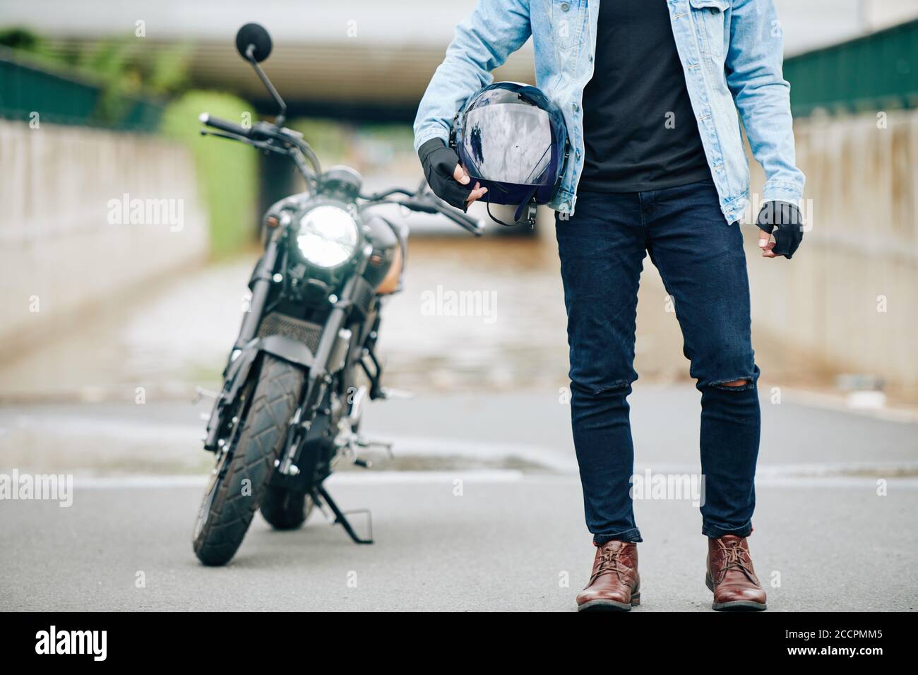 Cropped image of motorcyclist in jeans and leather boots holding helmet,  his motorcycle is in background Stock Photo - Alamy