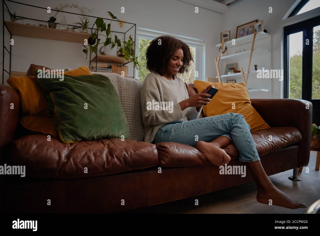 Smiling african woman sitting on comfortable couch using smartphone at ...