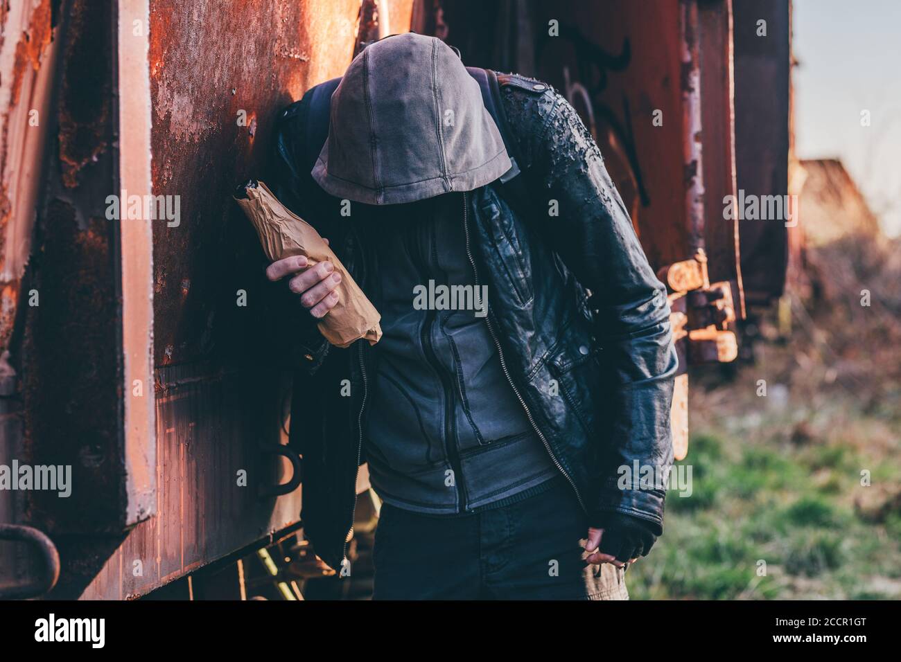 Homeless drunkard drinking alcohol next to old train wagon, drunk man with hoodie in alcoholism concept, selective focus Stock Photo