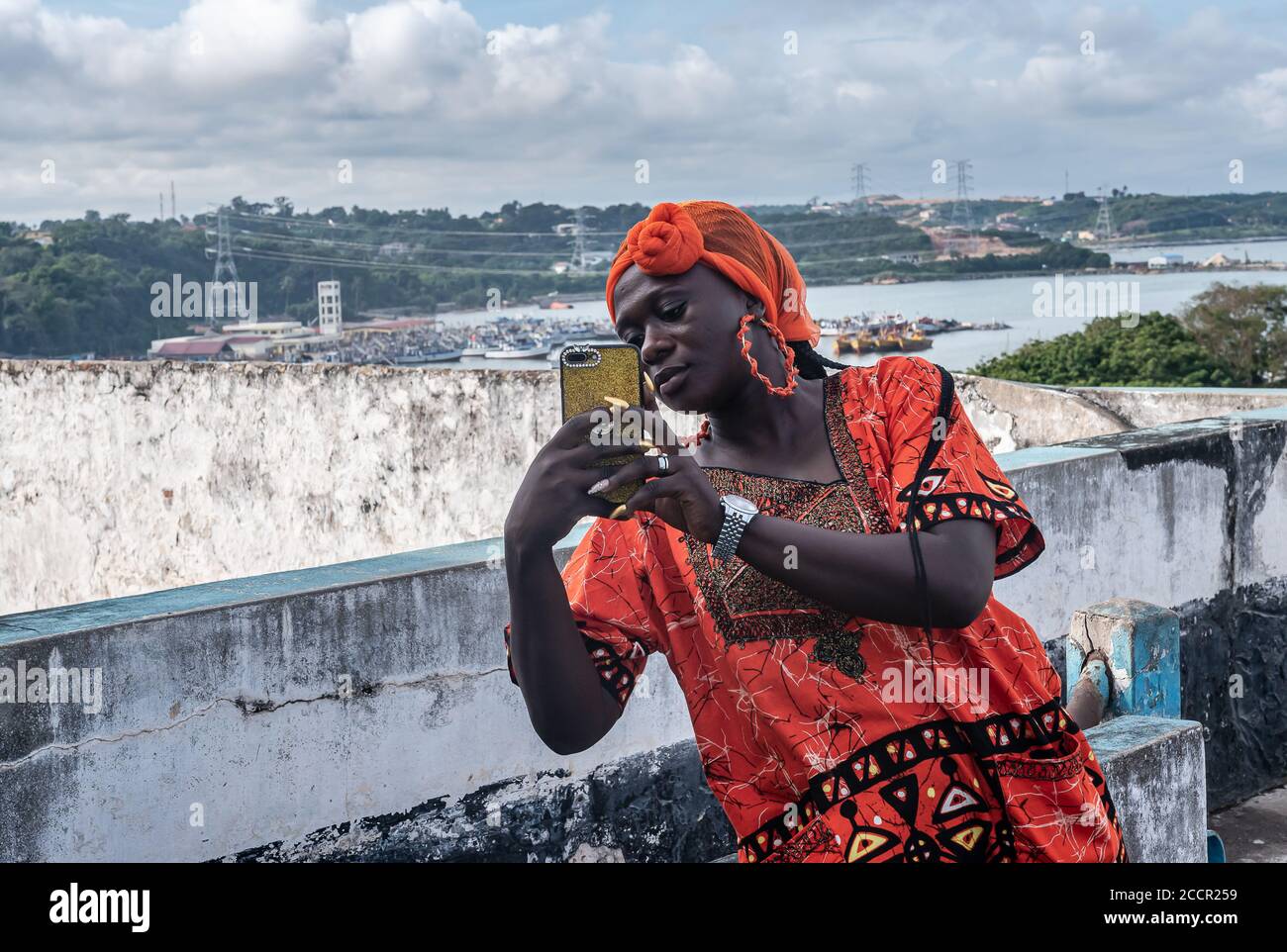 African woman with a mobile phone in hand and wearing traditional orange african dress. The place is Takoradi Ghana West Africa. Stock Photo
