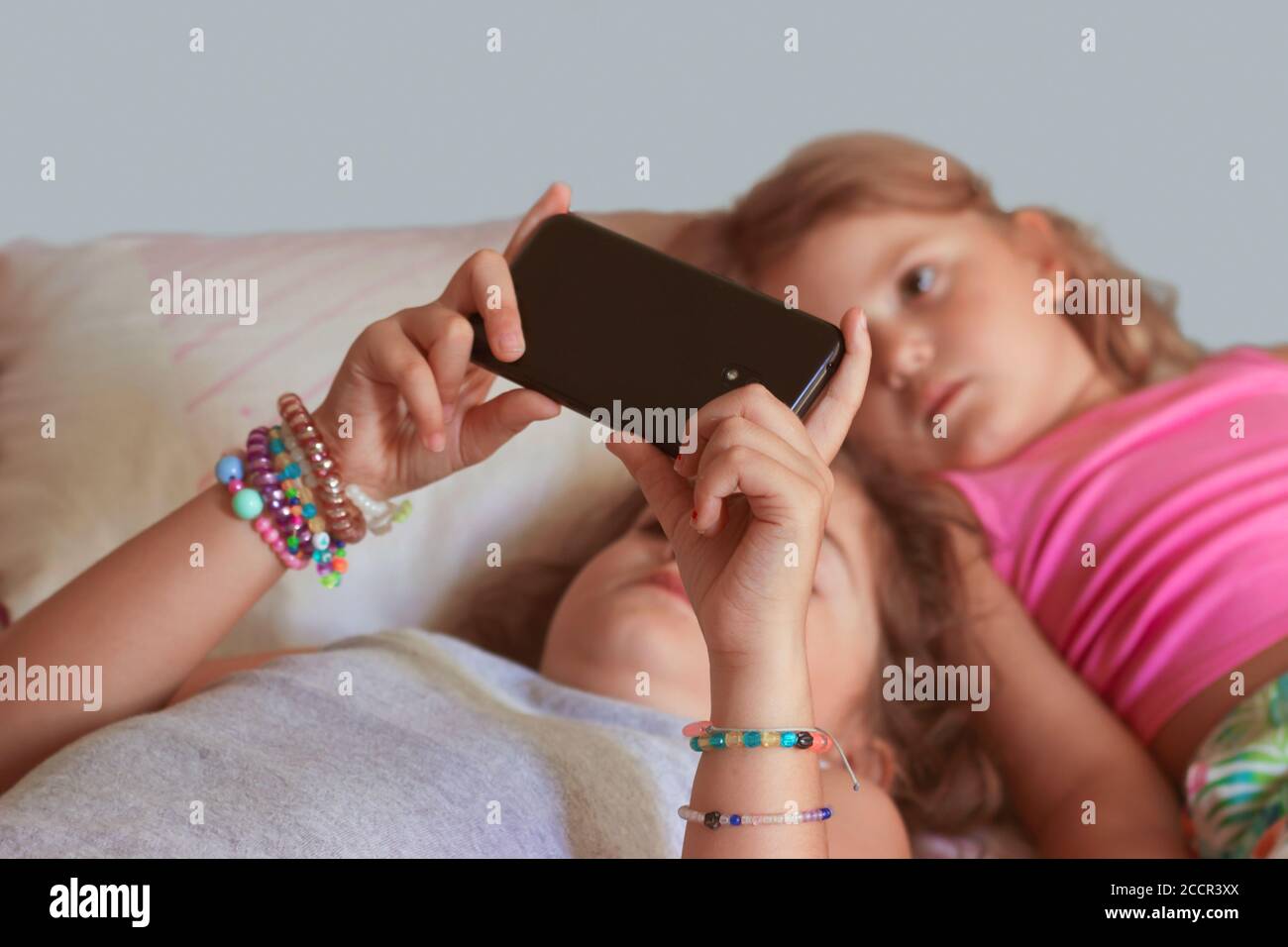 Two little girls, laying on the bed, watching videos on smartphone, being boring; Focus on smarthphone Stock Photo