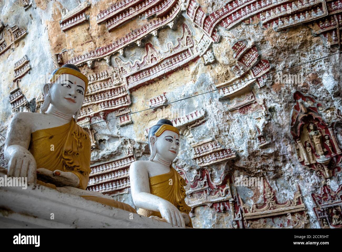 Small figurines carved on walls at Kaw Goon caves. Two bigger white and golden statues representing sitting Buddha at the foreground. Hpa An, Myanmar Stock Photo