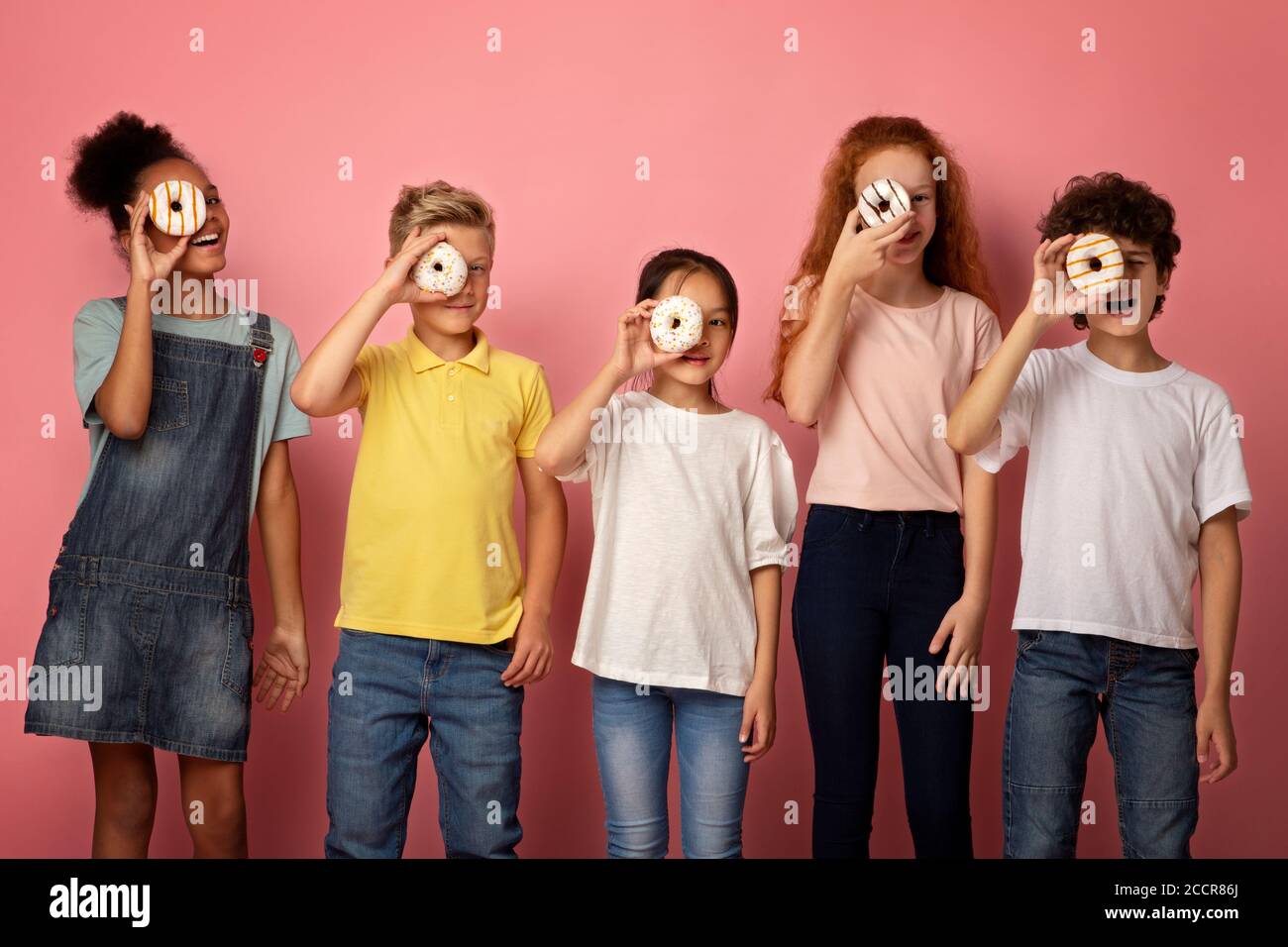 Diverse schoolkids holding yummy donuts in front of their faces over pink background Stock Photo