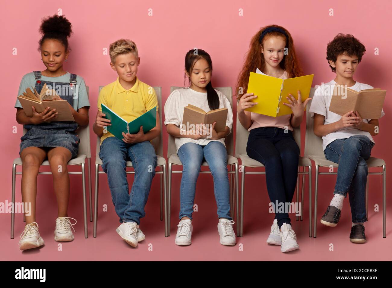 Diverse kids with textbooks and notebooks sitting on chairs, getting ready for school lesson on pink background Stock Photo