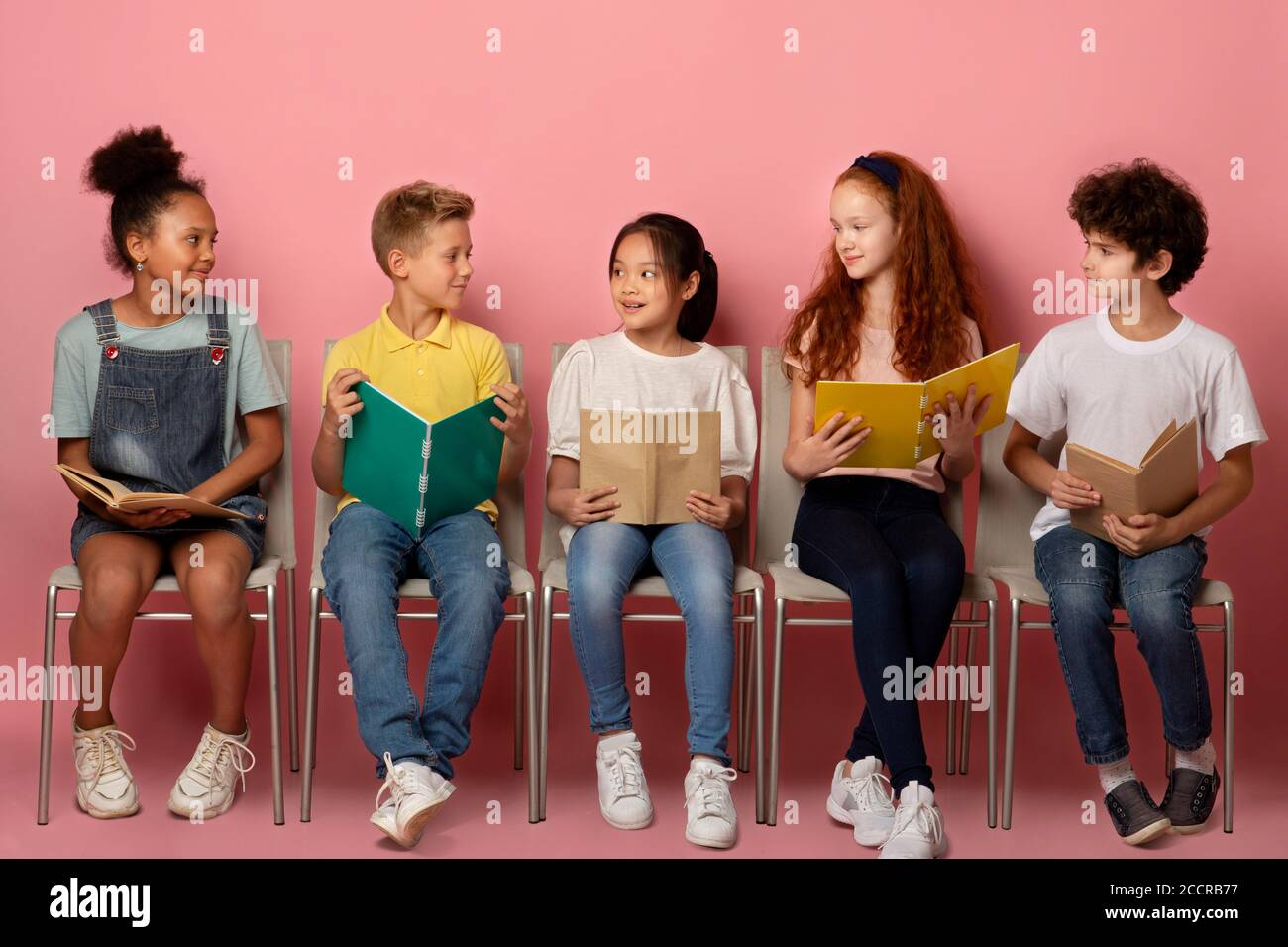 Cute multiracial schoolkids with study materials on chairs over pink studio background Stock Photo