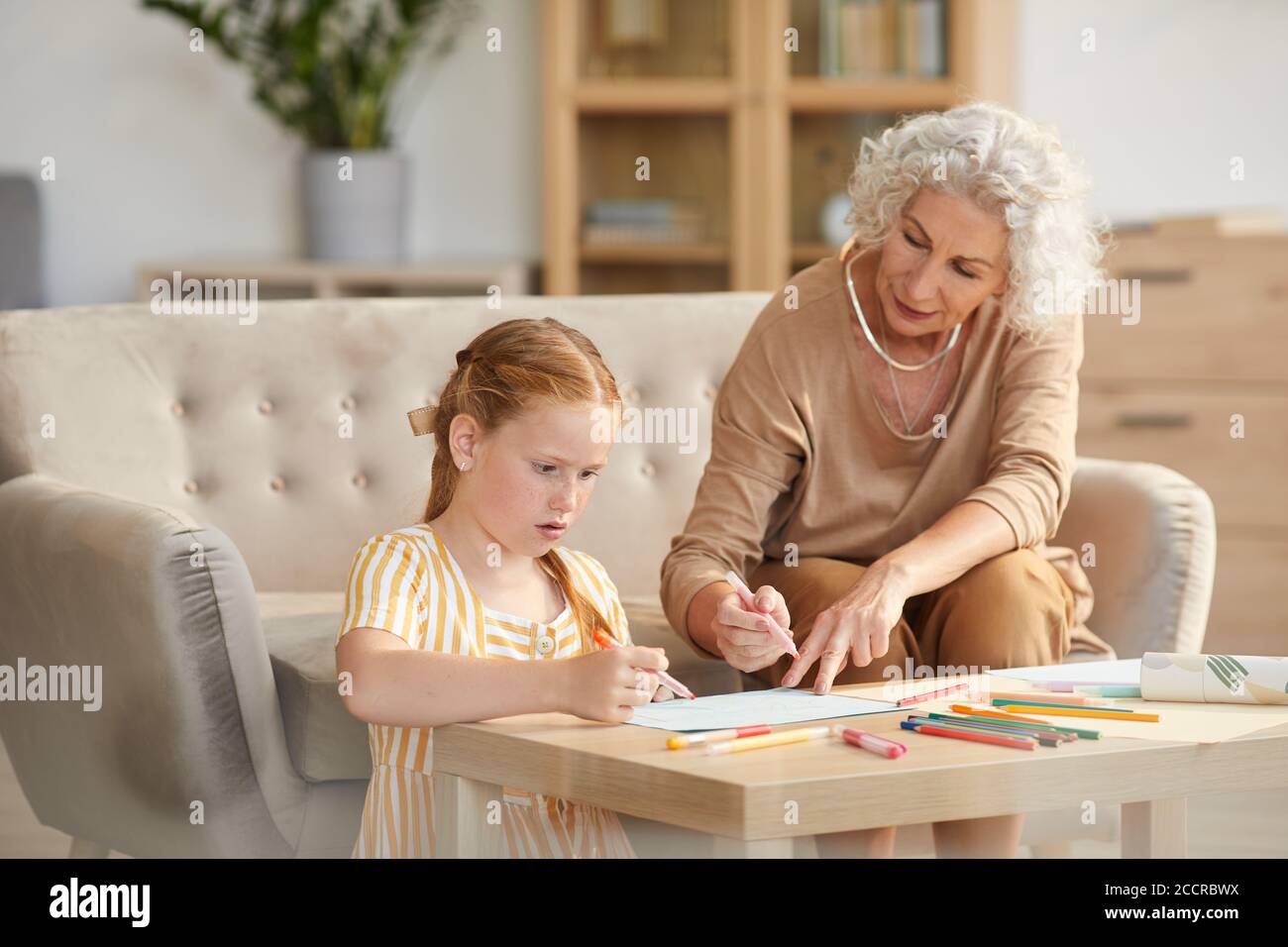 Warm toned portrait of senior woman babysitting cute red haired girl and drawing together while sitting in cozy living room Stock Photo