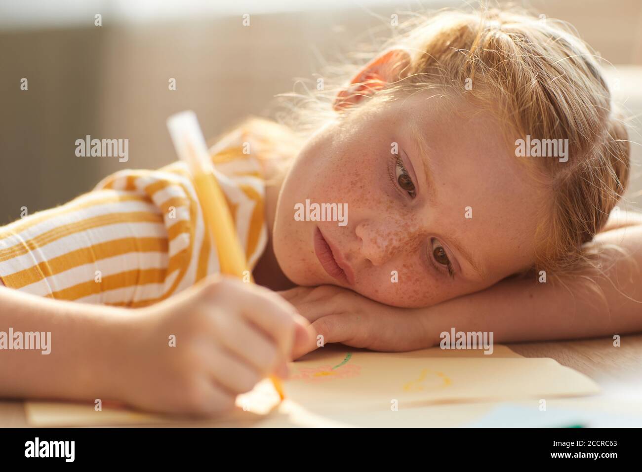 Warm toned portrait of cute red haired girl drawing pictures while lying her head on table in sunlight Stock Photo