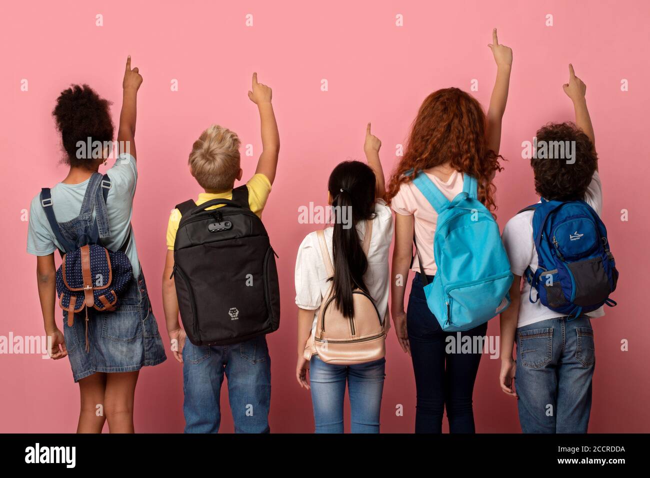 Back view of schoolkids with backpacks pointing up or touching virtual screen on pink background Stock Photo