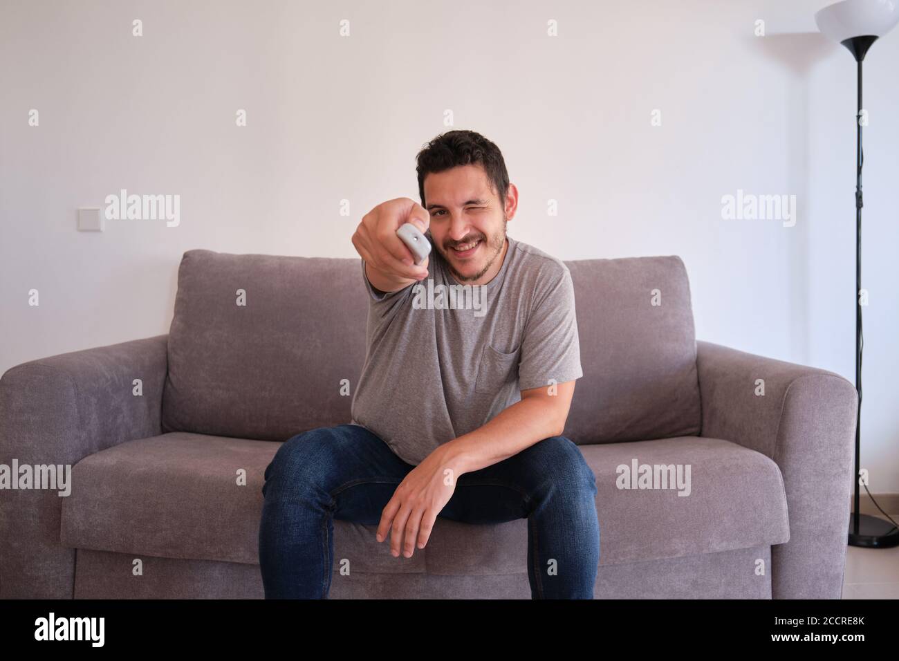Portrait of a smiling young man sitting on a sofa and changing TV channels with the remote control on his hands. Stock Photo