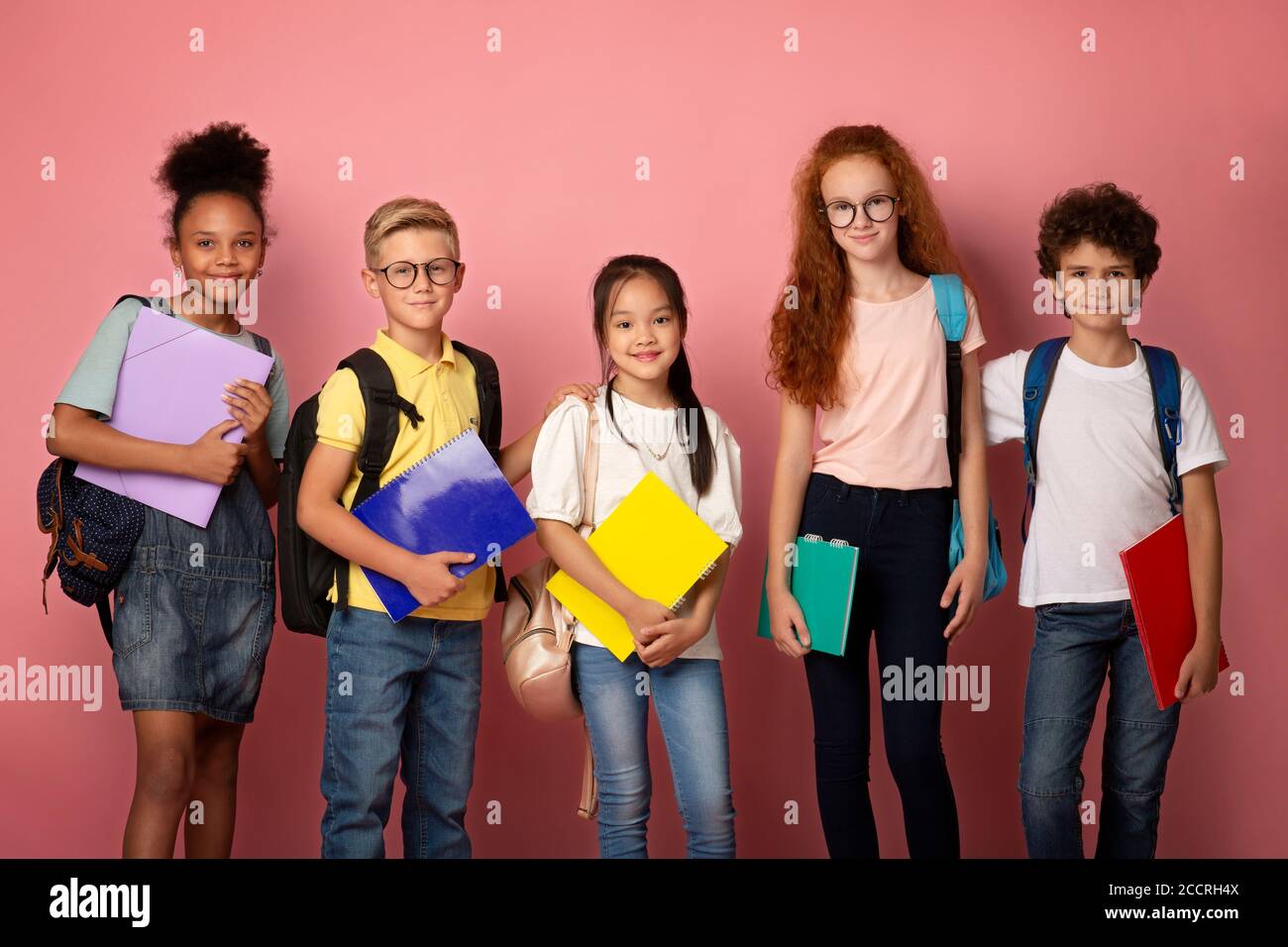 Schoolkids of different nationalities with notebooks and backpacks posing over pink background Stock Photo