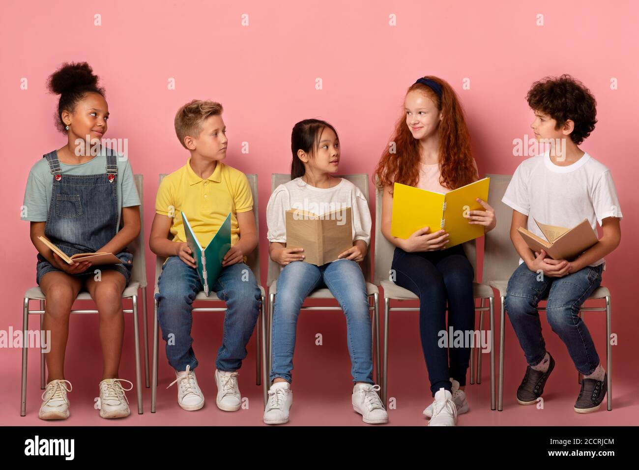 Classmates of different races with school supplies looking at each other on chairs over pink background Stock Photo