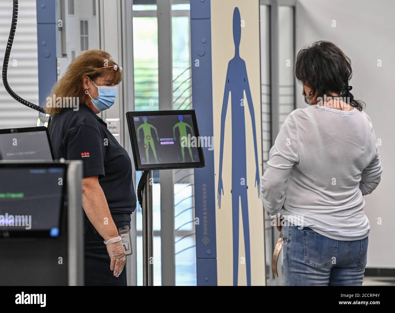 24 August 2020, Brandenburg, Schönefeld: A traveler (r) is checked at the  body scanner by a member of the security control staff in the building of  Terminal T5 at Berlin-Schönefeld Airport. On
