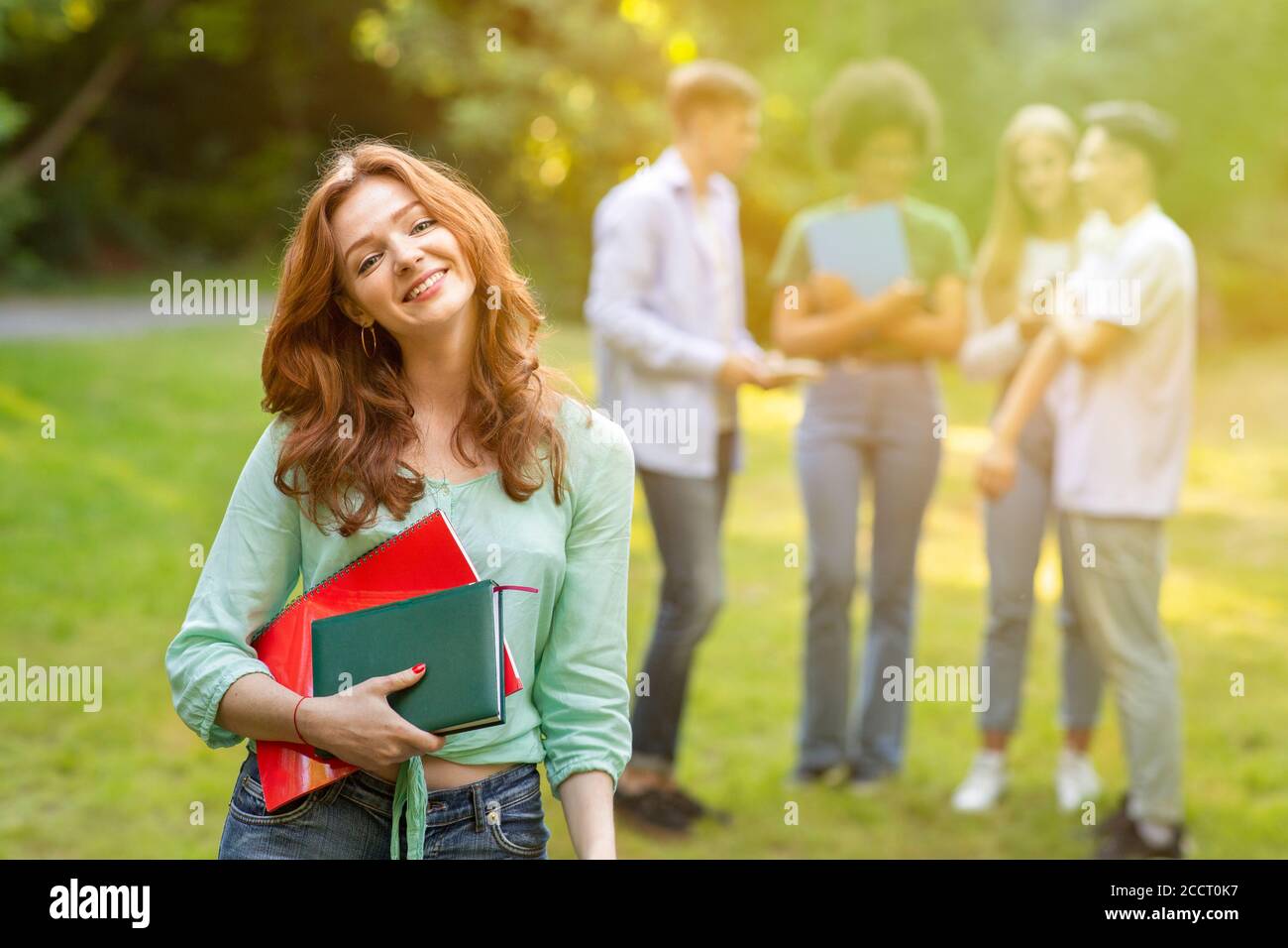 Cute student girl with workbooks posing outdoors with her classmates on background Stock Photo