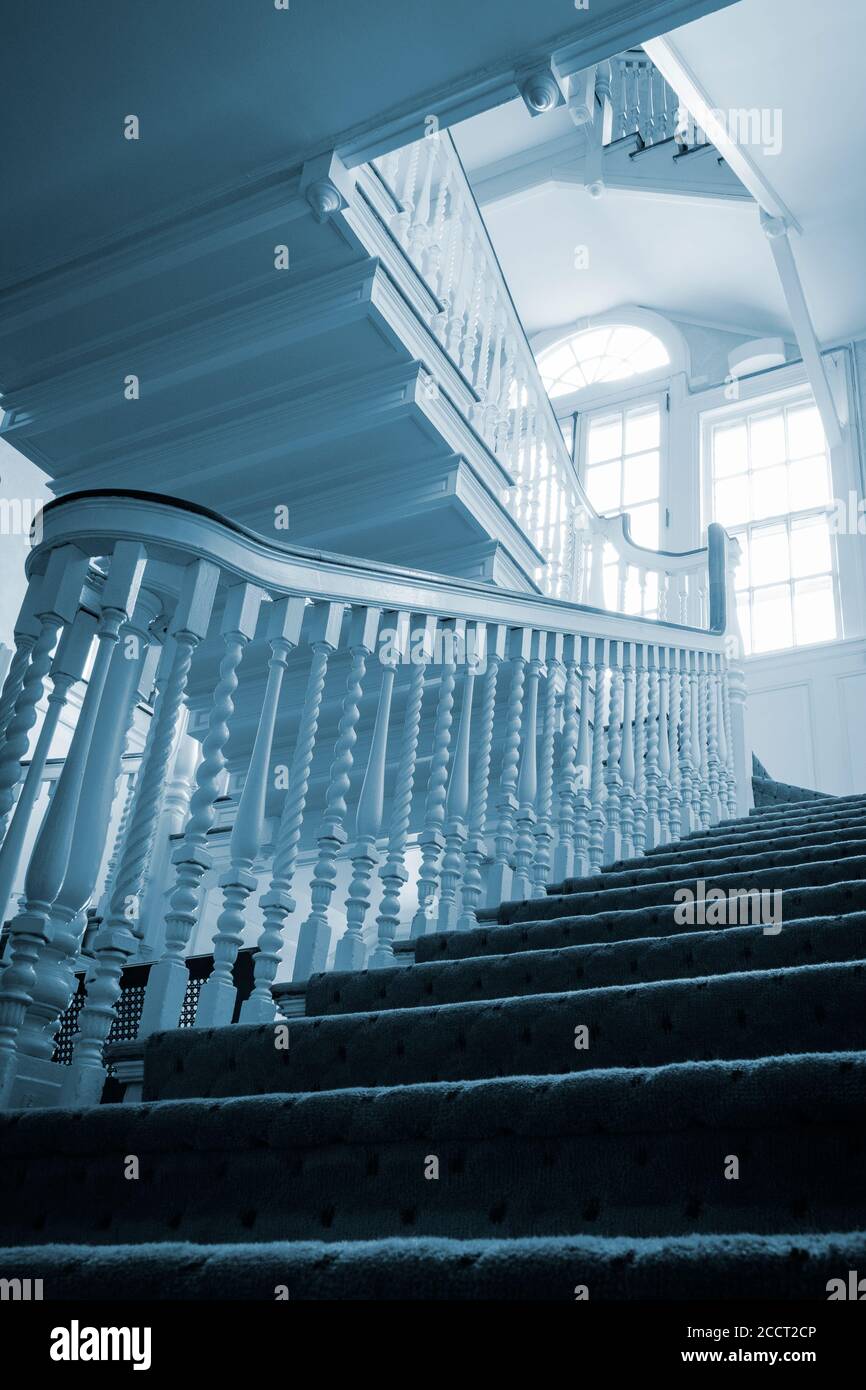Mysterious old wooden staircase leads upward past bright windows and blue cast daylight. Stock Photo