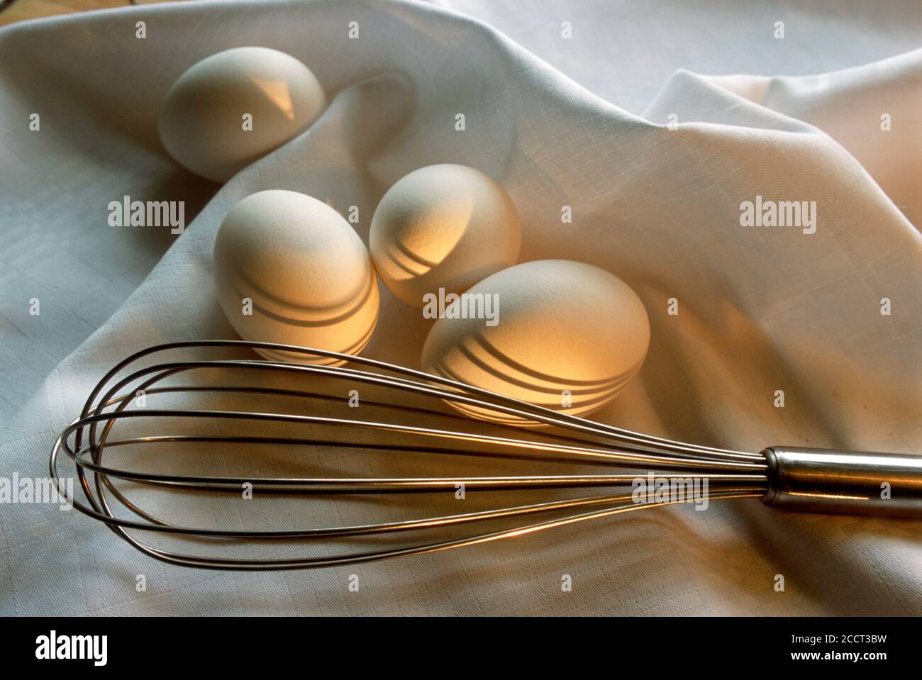Still life of whisk with chicken eggs on white cloth in kitchen Stock Photo