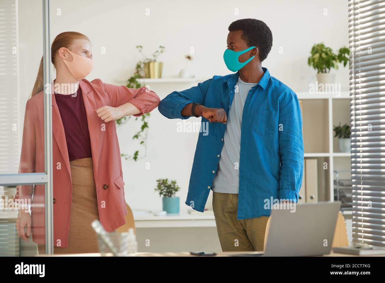 Portrait of young woman wearing mask bumping elbows with African-American colleague as contactless greeting while working in post pandemic office Stock Photo