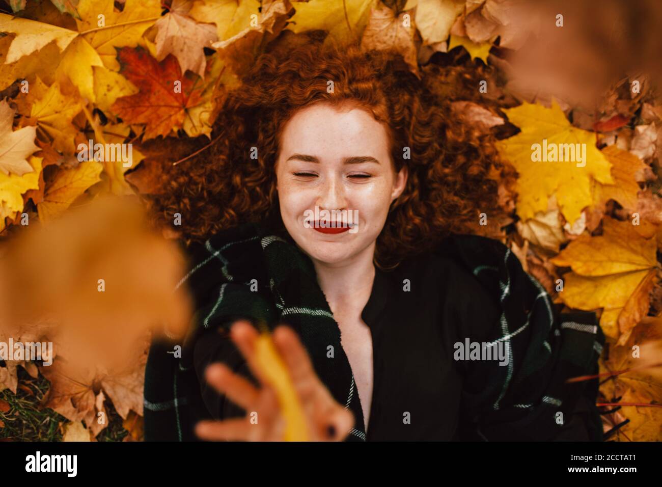 Young happy redhead woman lying on field with orange dry leaves in autumn Stock Photo
