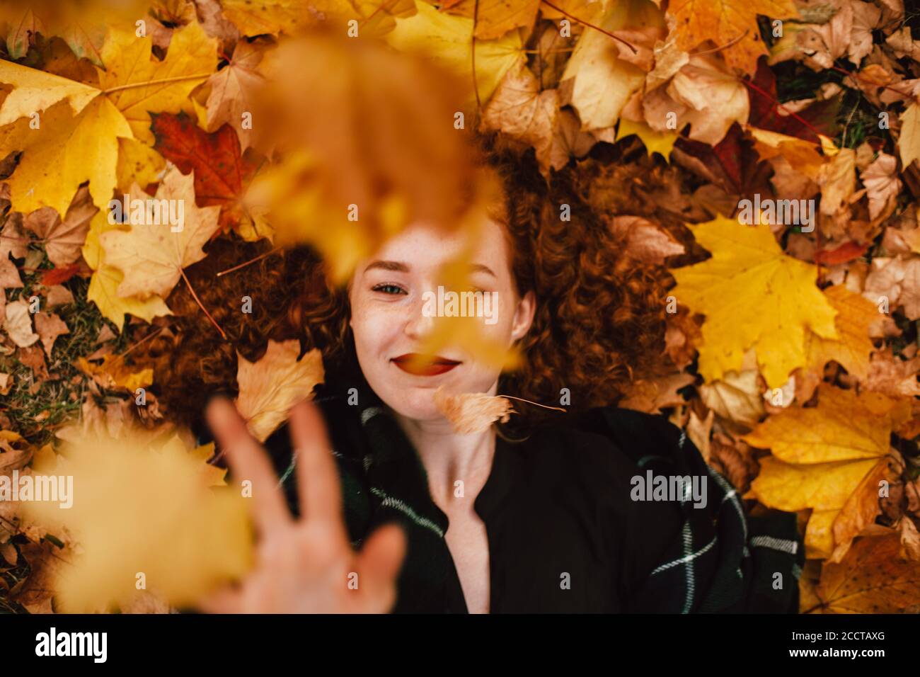 Young happy redhead woman lying on field with orange dry leaves in autumn Stock Photo