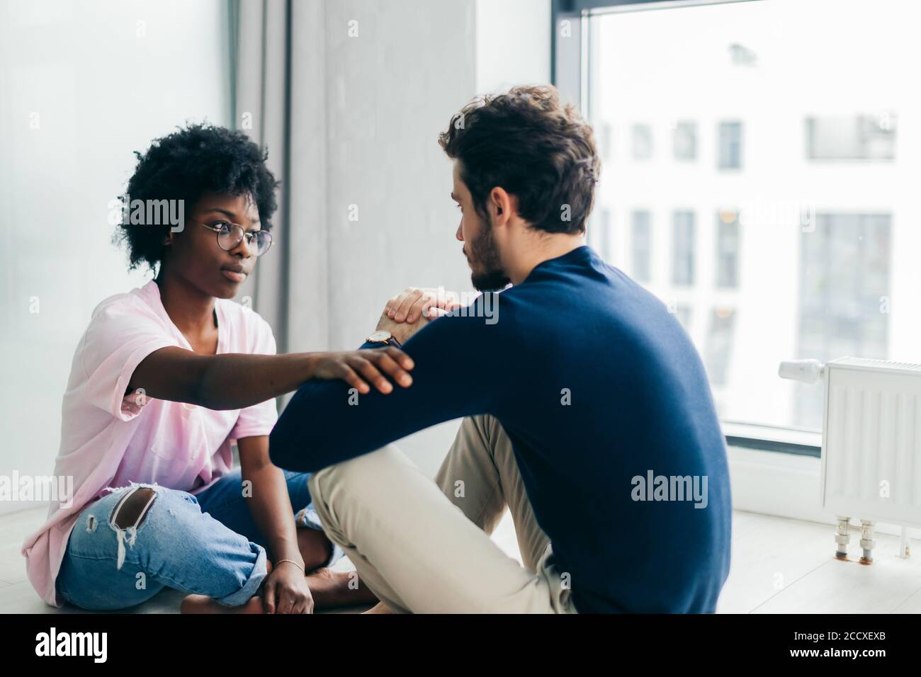 Young African woman and Caucasian man sitting near the window, staring at each other meditating together, free their minds from thoughts and worries. Stock Photo