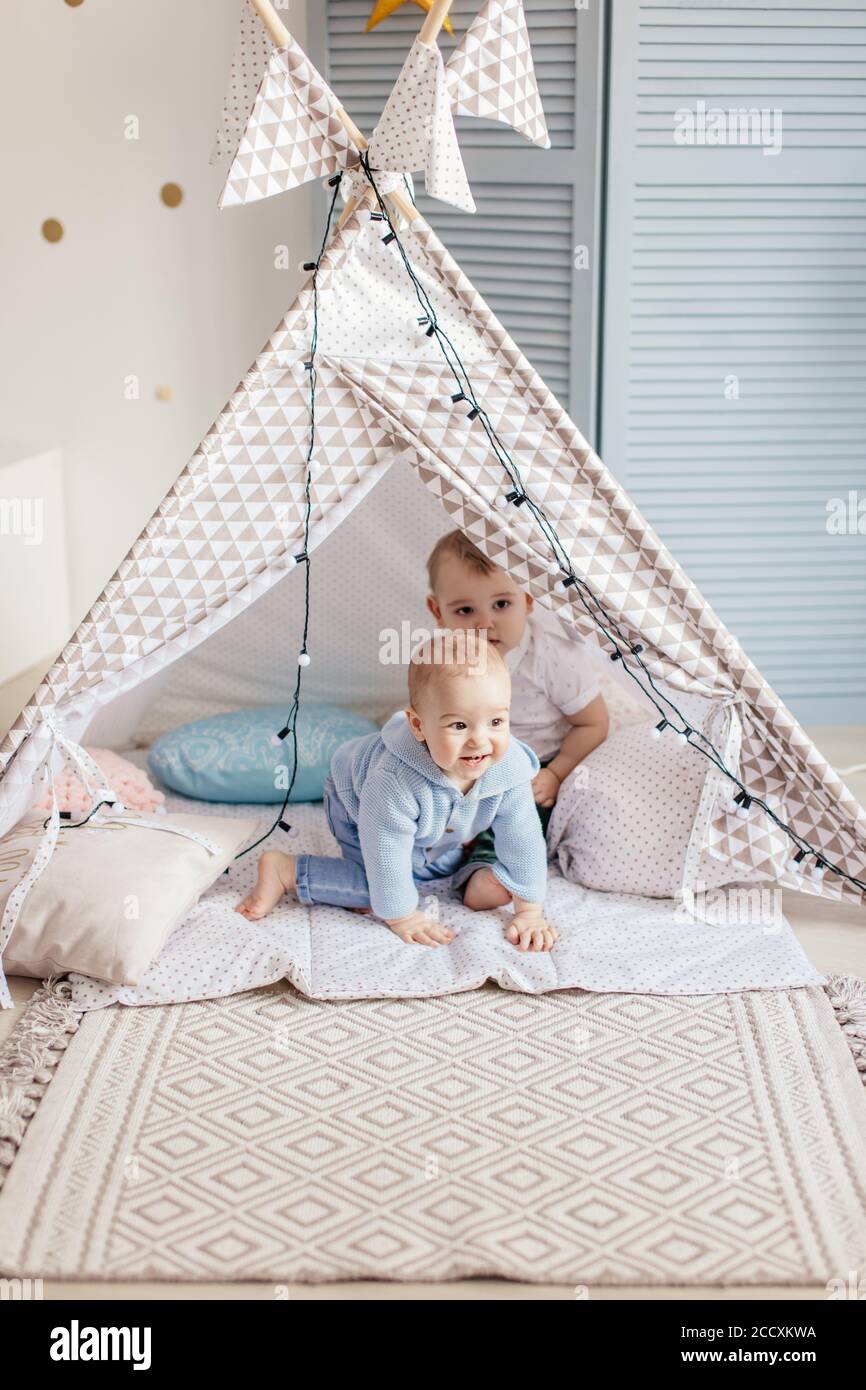 Two little infant male kids playing in toy tent indoor, in a pastel coloured playing in room, learning to smile, roll over, sit up, wave, clap, pick o Stock Photo