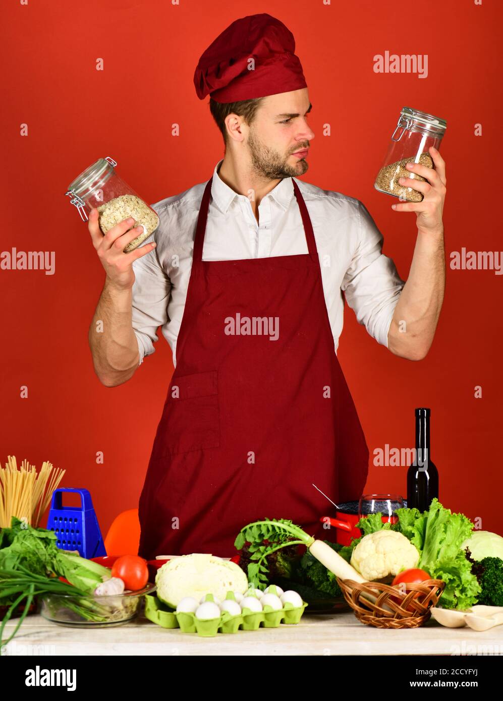 Chef with curious face holds jars with porridge on red background. Man in cook hat and apron uses groats. Cook works in kitchen near table with vegetables and tools. Cuisine and cooking concept. Stock Photo